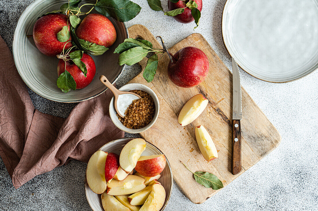 Top view of table setting with fresh red ripe apples in plates near cutting board with half cut pieces, cutlery and napkin on gray surface in daylight