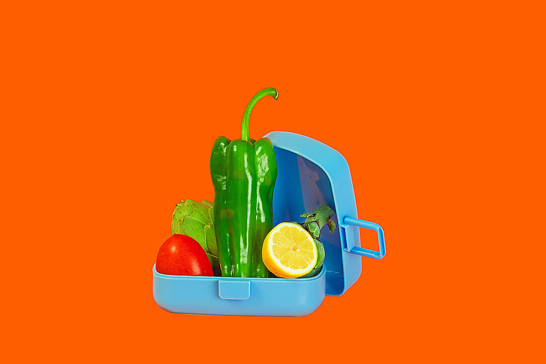Eine offene Brotdose, gefüllt mit leuchtend grünen Paprika, Salat, Tomaten und einer Zitronenscheibe vor einem kontrastierenden orangen Hintergrund