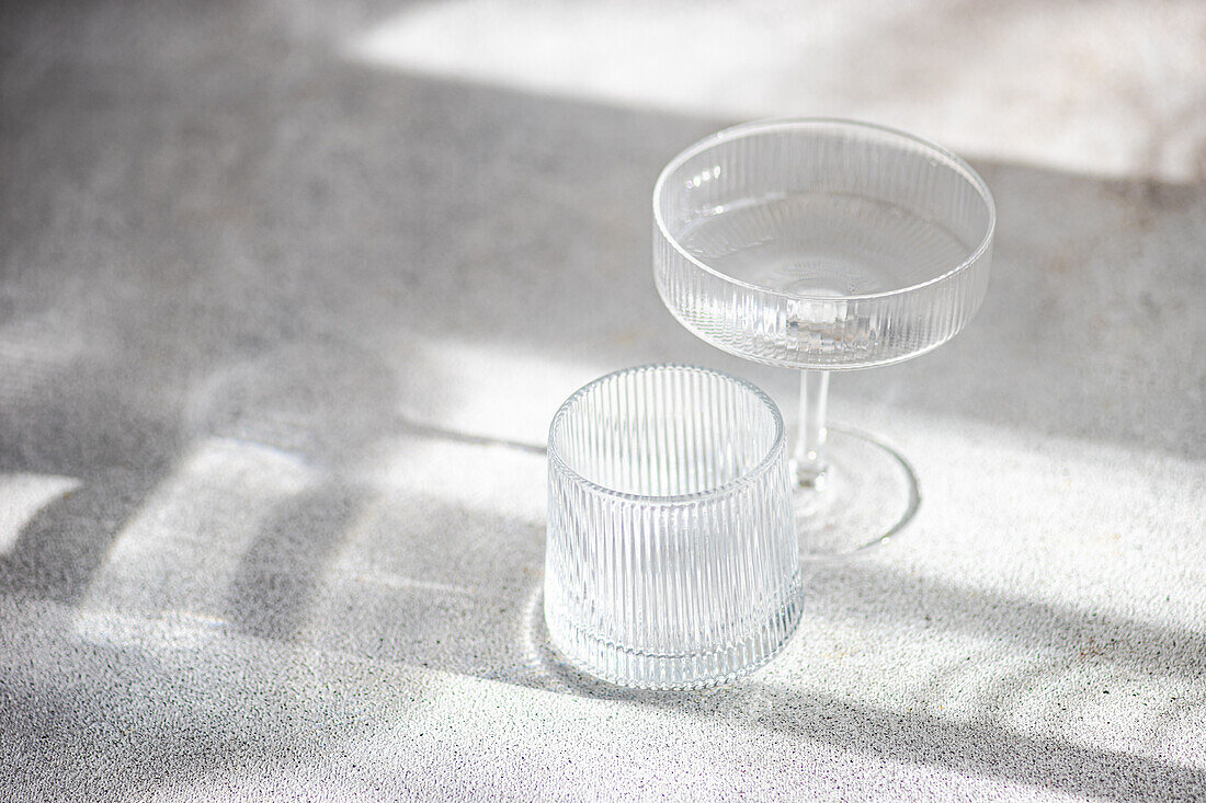 Zwei elegante Gläser, die in sanftes Sonnenlicht getaucht sind und ein Spiel aus Licht und Schatten auf einer strukturierten Oberfläche erzeugen