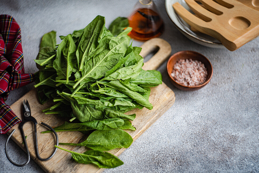 Vibrant spinach leaves placed on a wooden board, accompanied by a bowl of pink salt, a bottle of oil, wooden spatula, and rustic scissors on a textured backdrop