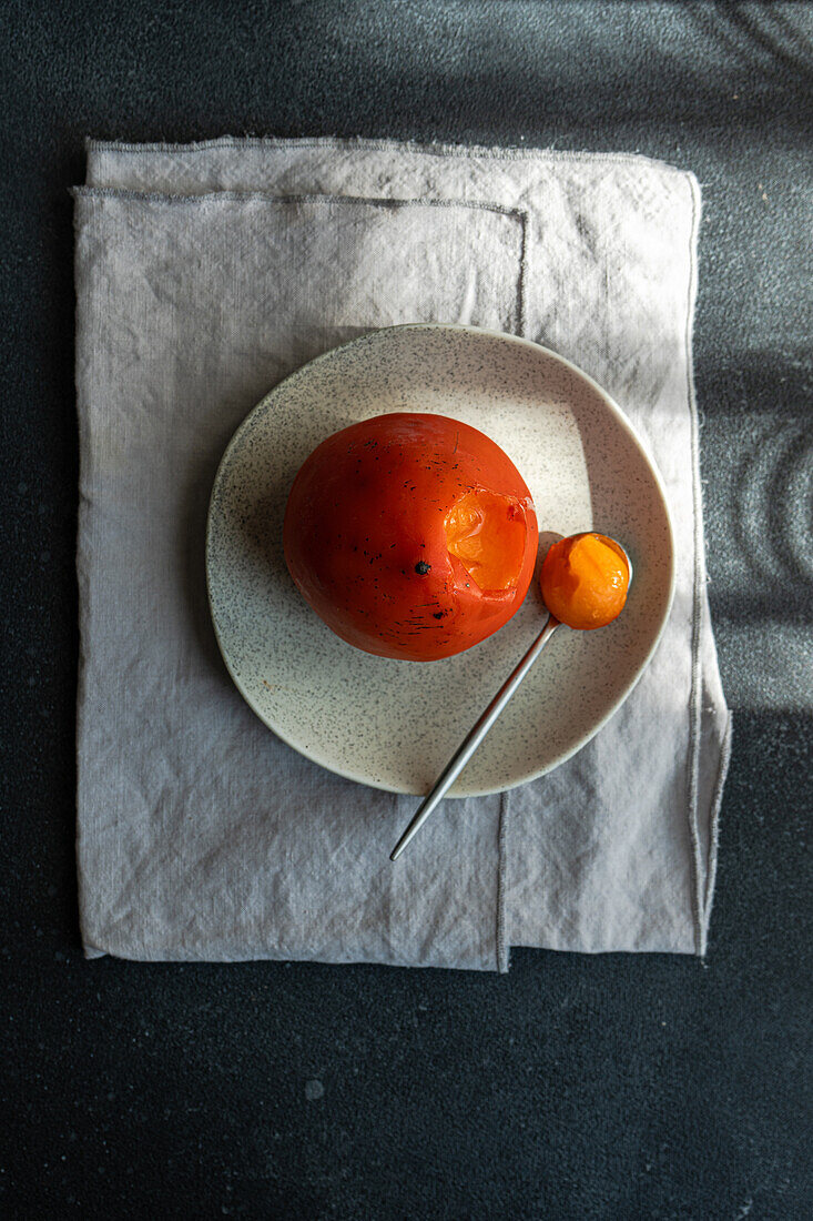 A vibrant, ripe persimmon rests on a ceramic plate beside a spoon, with dramatic shadows accentuating its curves.