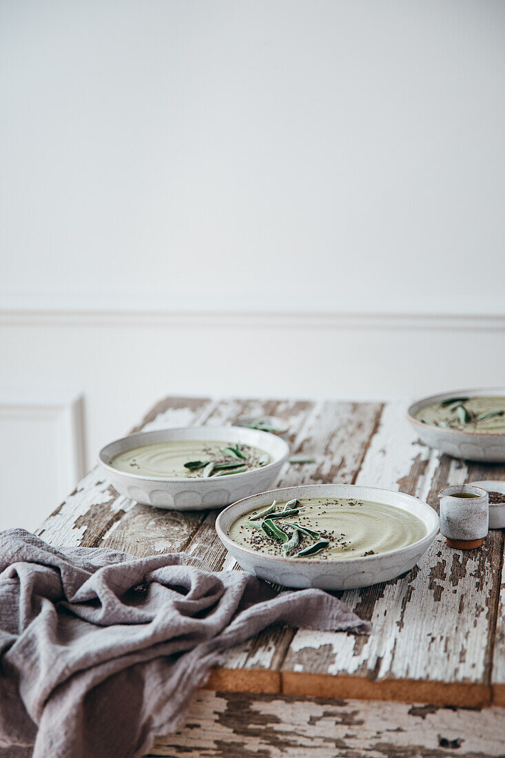 Ceramic bowls of green cream soup with sage served with seeds and herbs on wooden table with shabby surface against white background