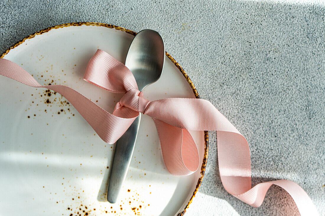 A top view of a shiny spoon tied with a soft pink ribbon on a ceramic plate with a golden rim, set against a textured background