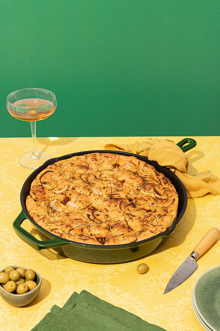 Flache, im Ofen gebackene italienische Focaccia in einer Pfanne, serviert mit frischen grünen Oliven und Honig in Schalen auf gelber Oberfläche