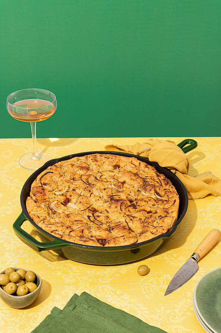 Flat leavened oven-baked Italian focaccia bread in pan served with fresh green olives and honey in bowls on yellow background surface