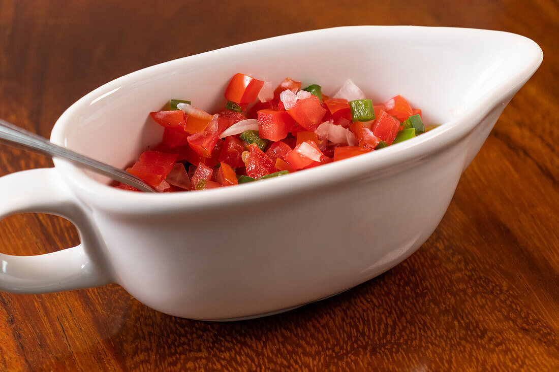 Bowl with spoon containing Pico de Gallo sauce prepared with slices of tomato onion green chili and placed on wooden surface in light