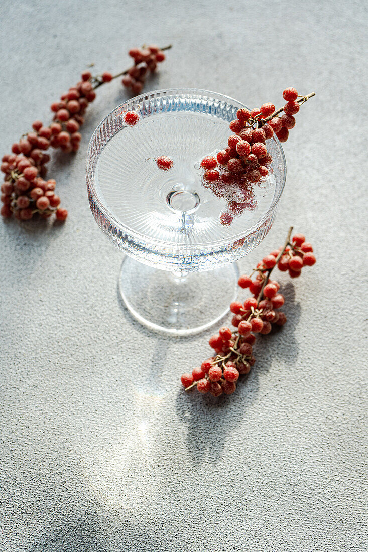 Ein Kuchenständer aus klarem Glas auf einer strukturierten Oberfläche, geschmückt mit kleinen roten Beeren, die um ihn herum verstreut sind