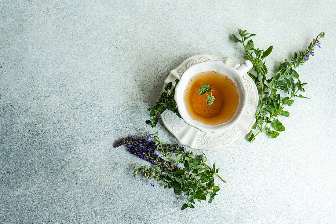 Draufsicht auf ein Glas Blumentee mit frischen Kräutern, serviert auf einem Keramikteller vor grauem Hintergrund