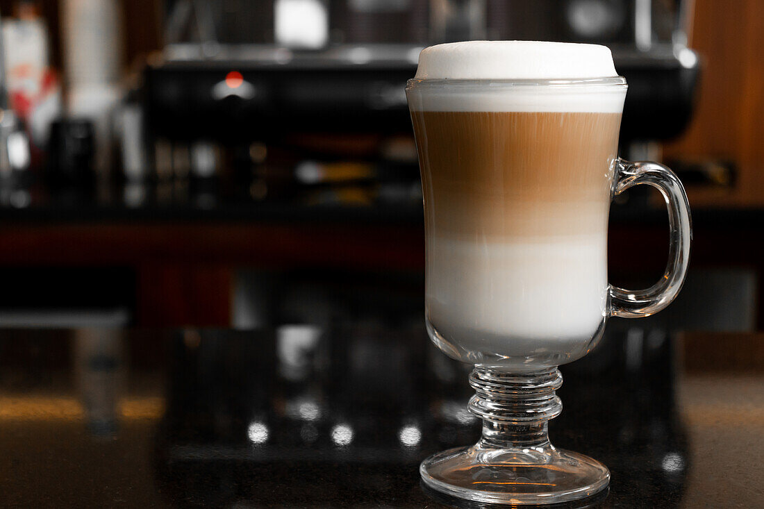 Ein Glas aromatischer heißer Cappuccino steht auf einem Tisch neben einer Kaffeemaschine vor einem unscharfen Hintergrund
