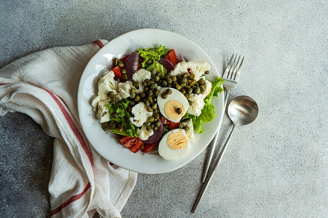 Draufsicht auf einen Teller mit Keto-Salat mit Salatblättern, gekochten Eiern, Kapern, Tomaten, roten Zwiebeln und Blumenkohlgemüse auf dem Tisch zwischen Serviette und Besteck