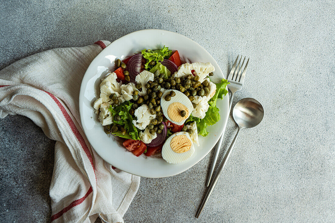 Top view of plate of Keto salad with salad leaves, boiled eggs, capers, tomato, red onion and cauliflower vegetables placed on table between napkin and cutlery