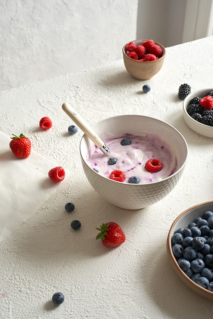 Hochformatige Schale mit Naturjoghurt und einigen frischen Früchten: Himbeeren, Heidelbeeren, Erdbeeren und Brombeeren neben Tellern mit frischen Früchten im Tageslicht