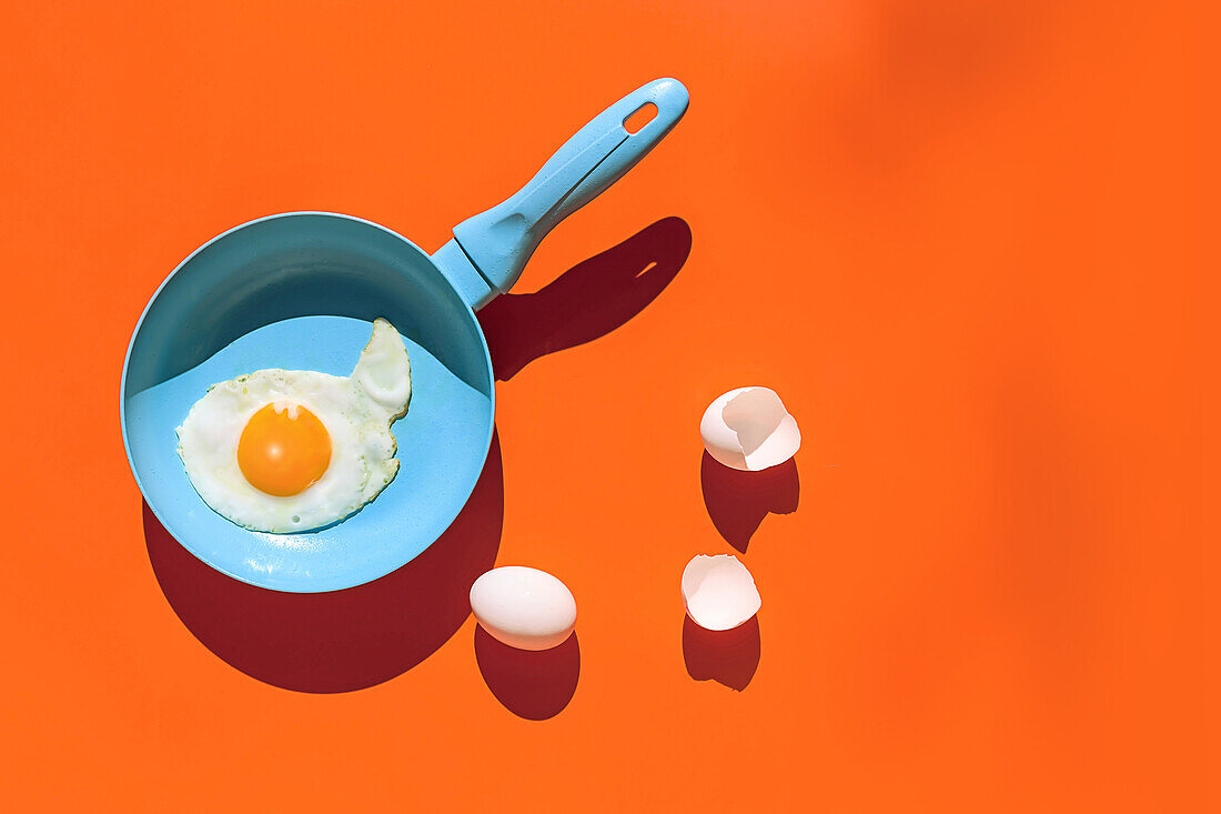 Top view of fried egg on blue frying pan near raw whole egg and eggshells with shadow isolated on orange background