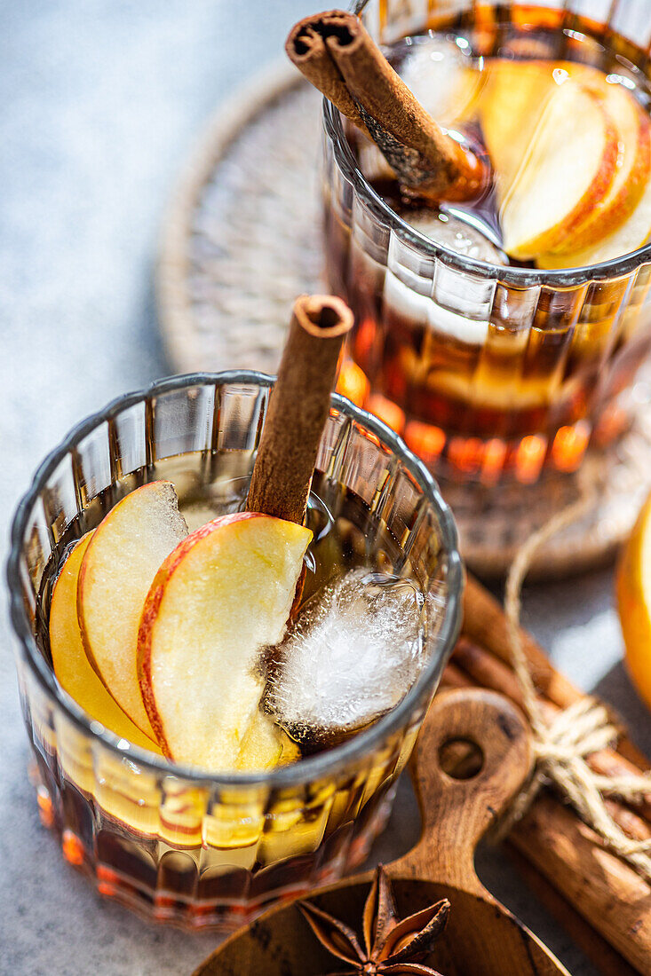 Fresh apple cider cocktails garnished with cinnamon sticks, star anise, and apple slices in a crystal glass filled with ice cubes on a textured background