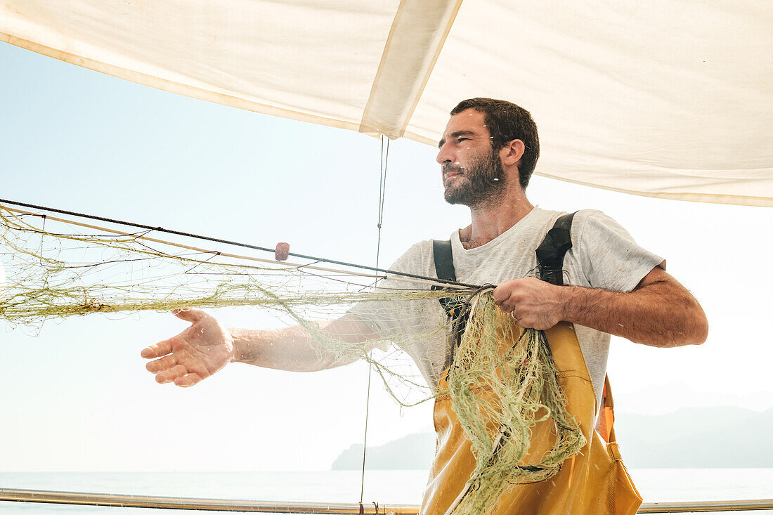 Fokussierter, bärtiger, männlicher Fischer in Uniform, der mit einem Netz auf Fischfang geht, während er auf einem Schoner in Soller nahe der Baleareninsel Mallorca arbeitet
