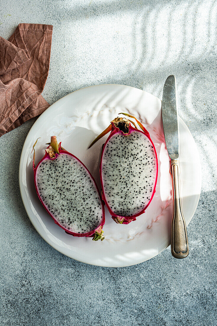 Two vibrant dragonfruit halves lie on a stylish white plate next to a vintage silver knife, with shadow patterns on a textured table.