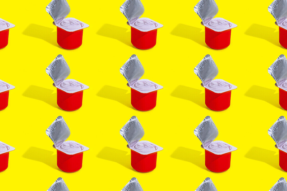 A repetitive pattern of red cups of strawberry yogurt, each with its foil lid opened, arranged neatly on a bright yellow background
