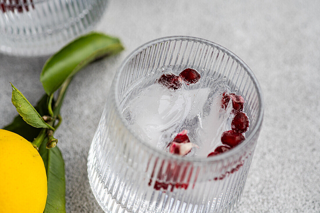 Close-up of a gin tonic glass with ice cubes, garnished with lemon and pomegranate seeds, served on a textured gray surface.