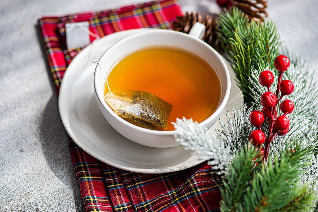 High angle of Christmas cup of tea placed on red napkin near holly and pine cones on gray table