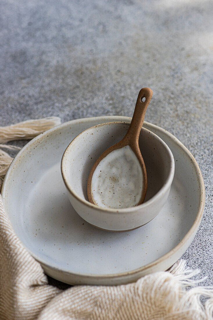 Hoher Winkel eines Keramikgeschirrsets, bestehend aus Schüssel und Teller mit Holzlöffel, neben einer Serviette auf einer grauen Fläche vor einem unscharfen Hintergrund