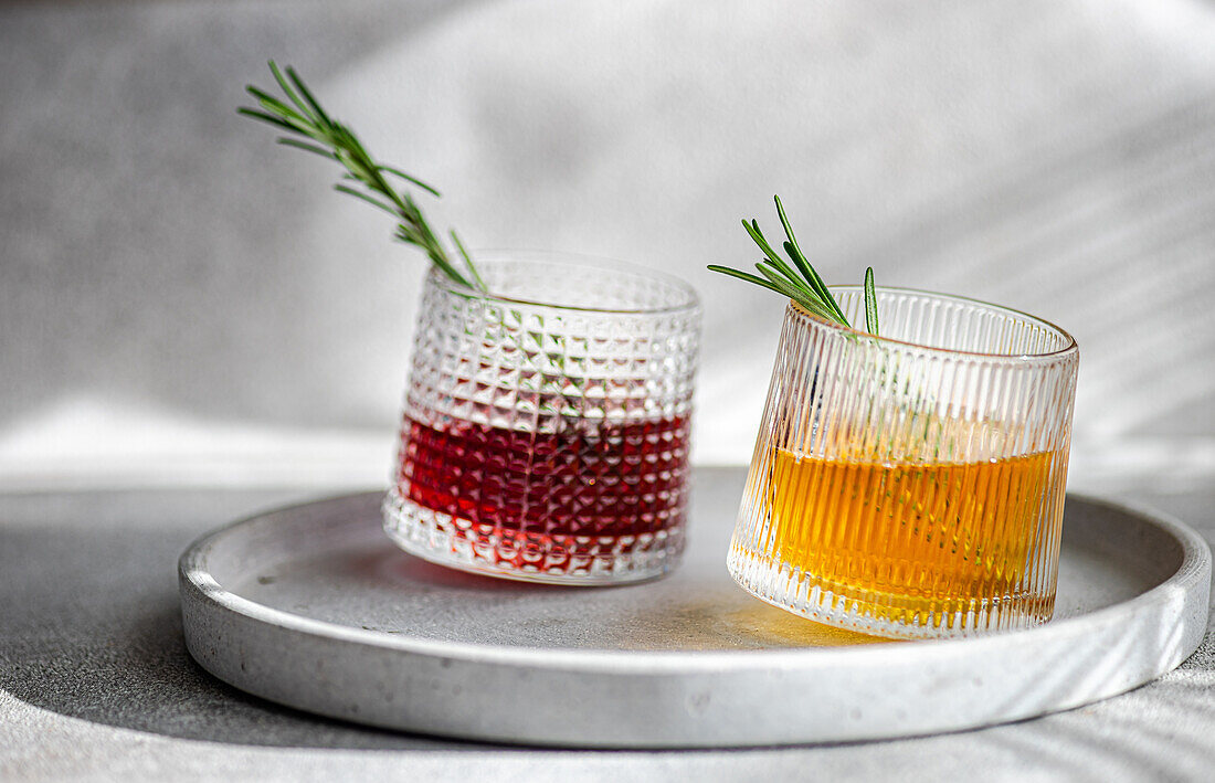 Zwei Cocktails in strukturierten Gläsern, einer mit Wodka mit Kirsch- und Apfelsaft, der andere mit einem goldenen Farbton, beide garniert mit Rosmarinzweigen auf einem runden Tablett