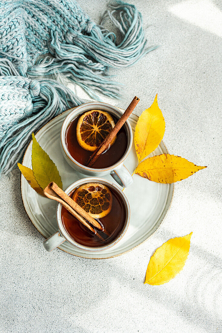 From above of cups of spiced tea adorned with cinnamon sticks, anise, and dried orange slices, nestled next to vibrant yellow leaves and a folded blue scarf on a soft gray backdrop