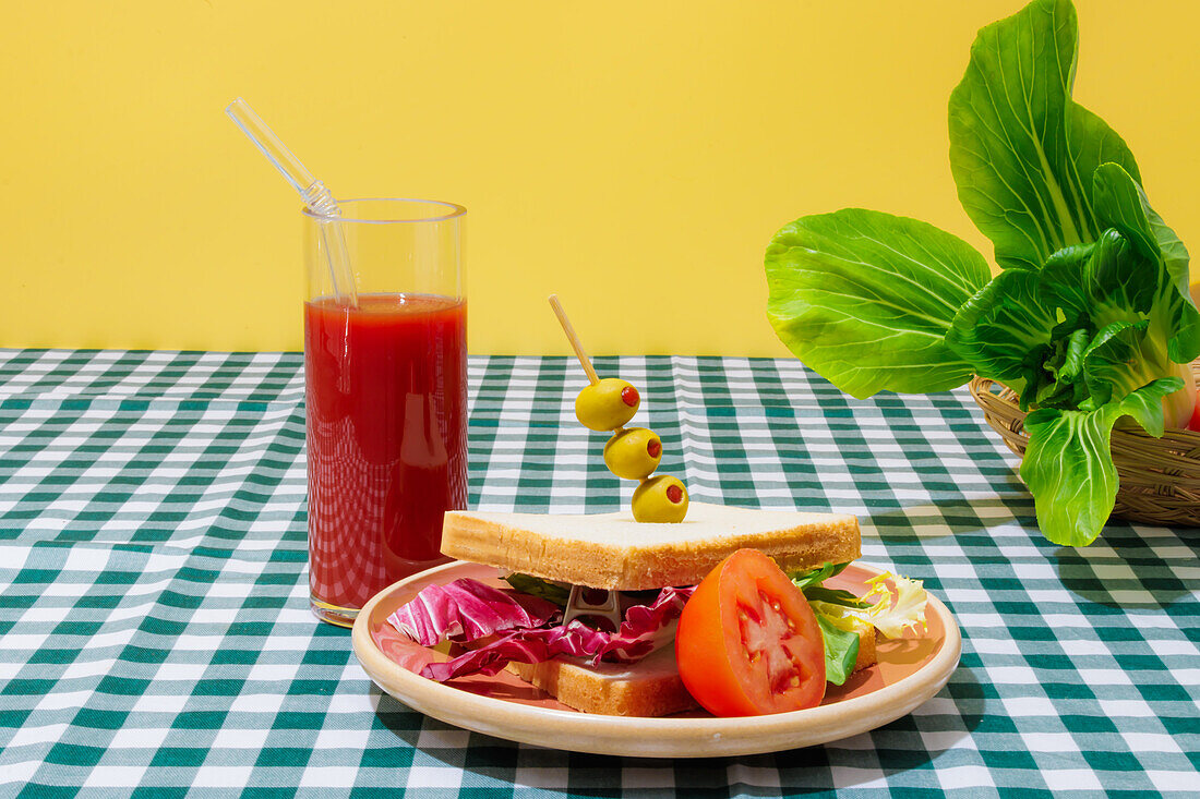 Appetitliches gesundes Sandwich mit frischem Salat, serviert auf einem Teller mit Oliven und einer halben Tomate neben einem Glas Erfrischungsgetränk mit Glasstrohhalm vor Salatblättern auf einem karierten Tischtuch