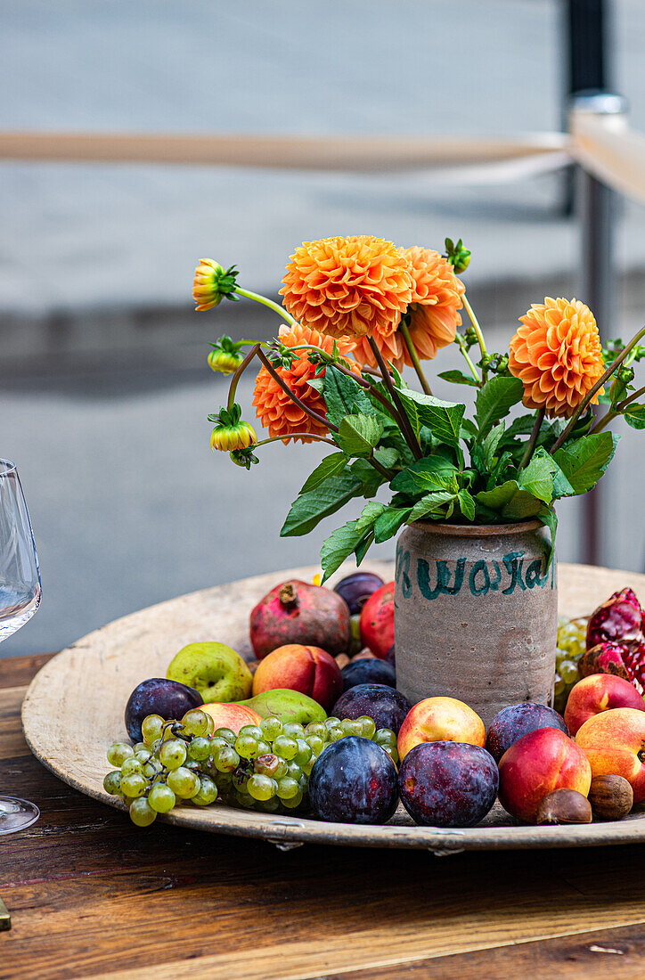 Bright orange dahlias in a rustic pot placed on a wooden table beside a platter of fresh assorted fruits.