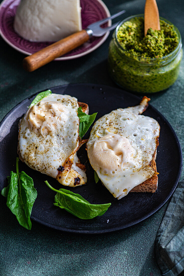 High angle of black plate with delicious homemade wholegrain toasts with fresh green spinach, fried egg and pesto sauce placed on gray table near plate with cheese