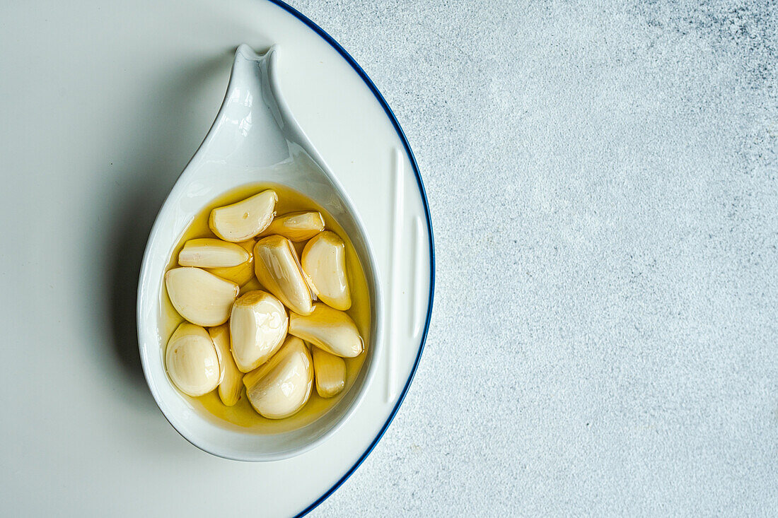 Top view of peeled and baked garlic cloves submerged in oil, showcased in a ceramic spoon placed on a white plate with a subtle blue border, against a light gray background