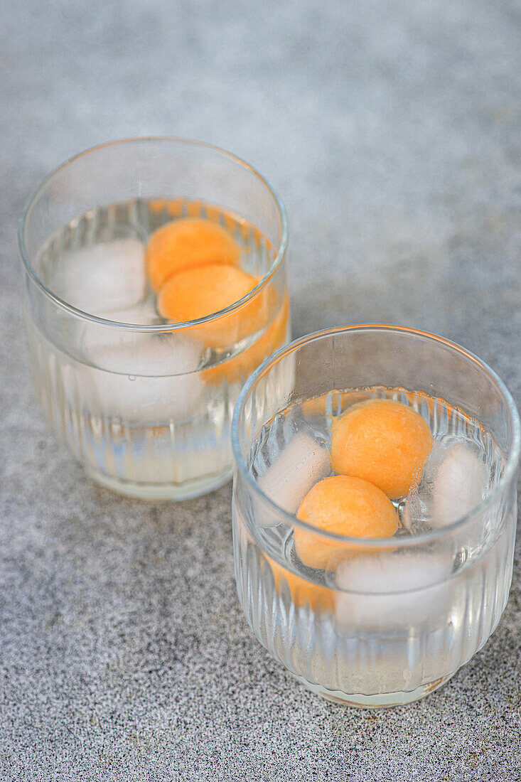 From above of pair of transparent glasses filled with melon spheres in iced beverage and placed on gray surface in daylight