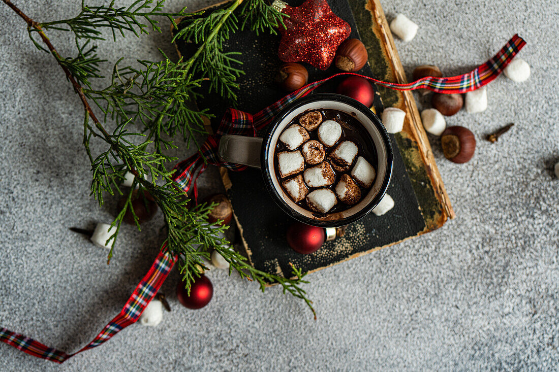 Draufsicht auf eine Tasse Kakao mit Marshmallow auf einem Buch neben Tannenzweigen auf grauem Hintergrund in der Weihnachtszeit