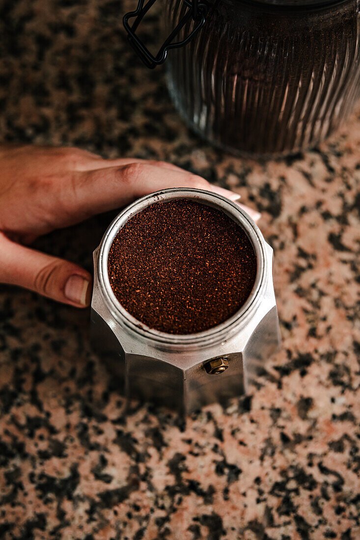 Top view of freshly ground coffee filled to the brim of stainless steel stovetop espresso maker with anonymous young person hand holding the base and a sealed coffee jar nearby on textured countertop