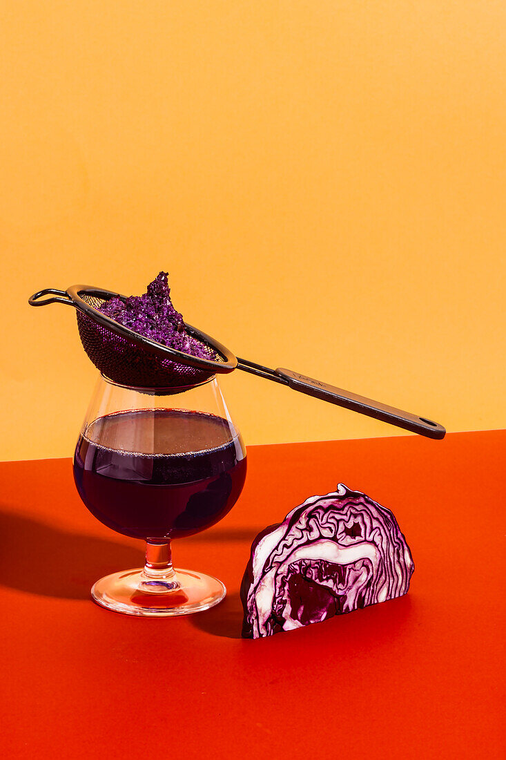 Healthy vegan antioxidant drink made with extracted red cabbage juice in glass with strainer on red and yellow background