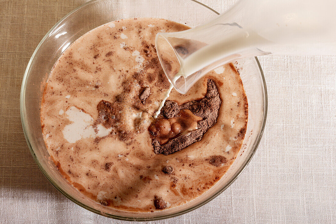 Top view of A stream of milk being poured into a glass bowl filled with chocolate placed on table