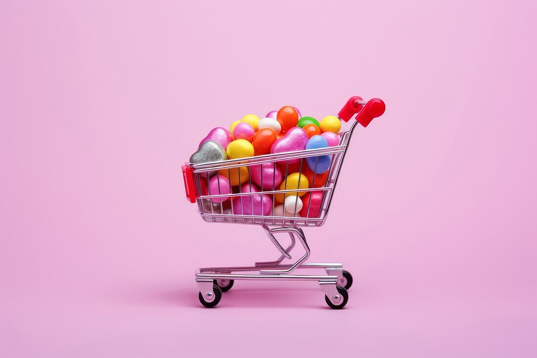 Komposition von Miniatur-Einkaufswagen mit verschiedenen bunten Süßigkeiten in der Nähe von gefallenen Süßigkeiten auf rosa Hintergrund platziert