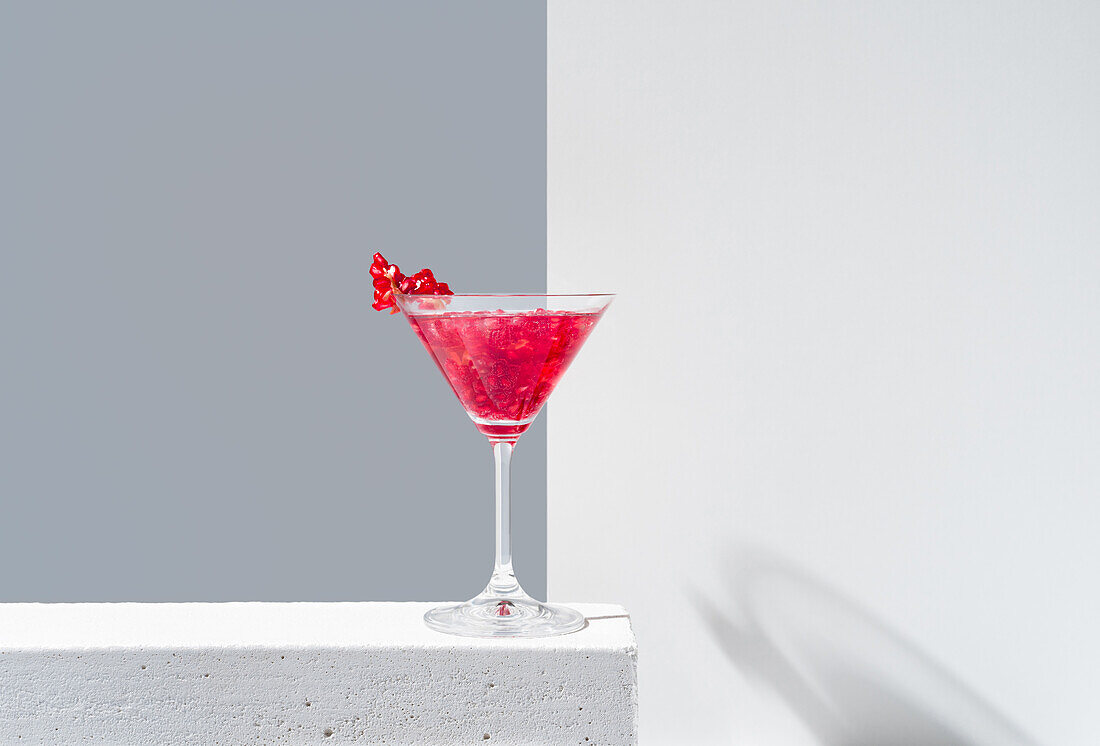 Glass filled with red pomegranate cocktail served with pomegranate seeds against a gray and white backdrop, casting shadow