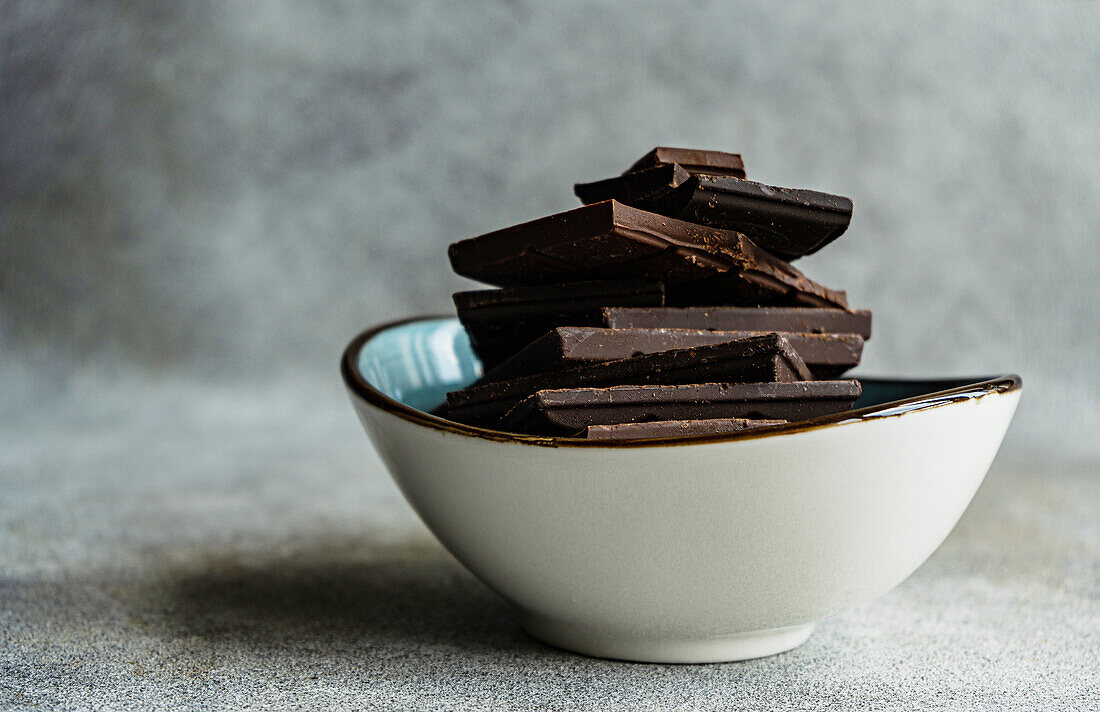 Pile of pieces of different kinds of chocolate placed in ceramic bowl on gray blurred table