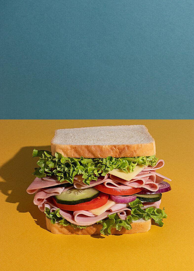 Leckeres Sandwich mit Schinken, Tomaten, Gurken- und Käsescheiben und frischem Salat auf blau-gelbem Hintergrund