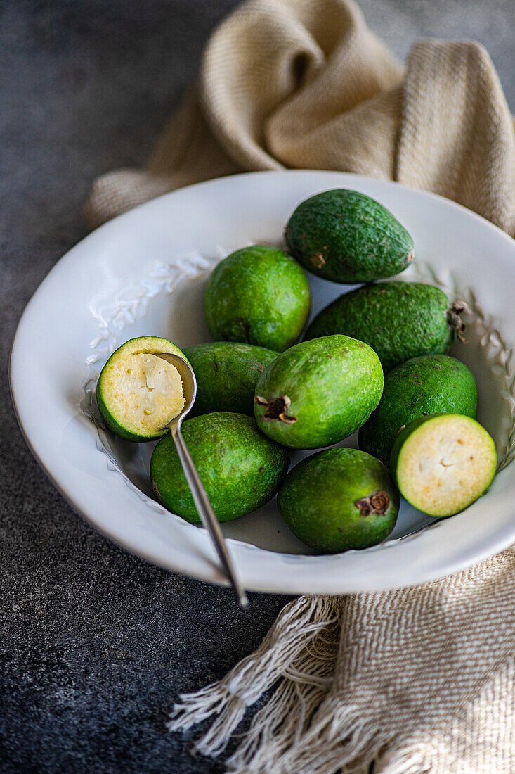 A composition of ripe feijoa fruits arranged in a white ceramic bowl, one cut open, with a textured napkin on a dark surface.