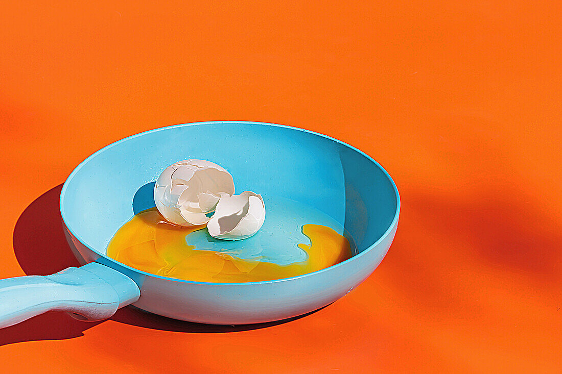 Blue frying pan with raw egg yolk and eggshell isolated on orange background