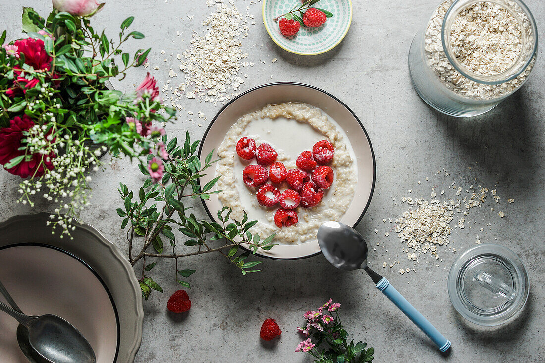 Bowl with porridge and heart shaped raspberries at grey concrete kitchen table with glass jar, branches, bowls and spoon. Preparing healthy vegan breakfast at home. Top view.