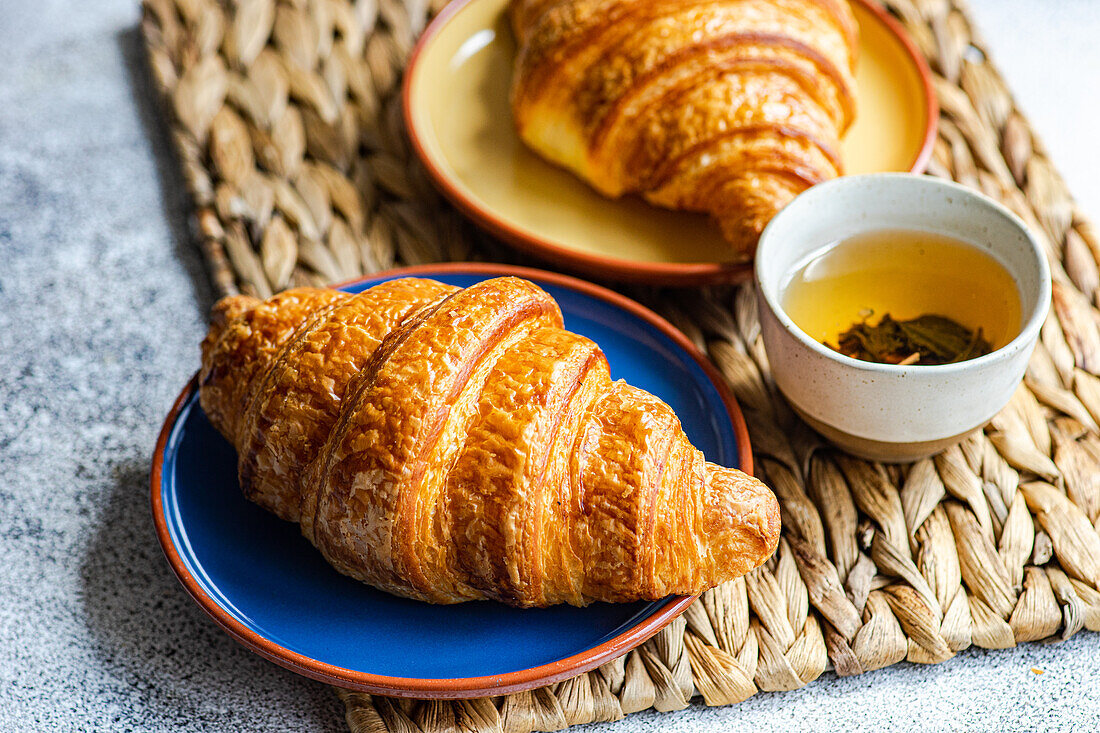 Nahaufnahme von frisch gebackenen Croissants auf bunten Keramiktellern auf einer braunen Serviette neben einer Tasse grünen Tees vor grauem Hintergrund