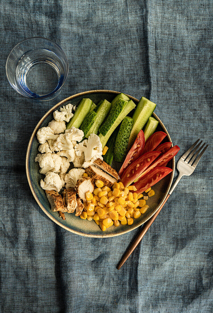 Top view of five ingredients bowl with raw organic vegetables cauliflower, tomato, cucumber, boiled sweet corn and barbeque chicken meat placed on gray napkin near fork and glass
