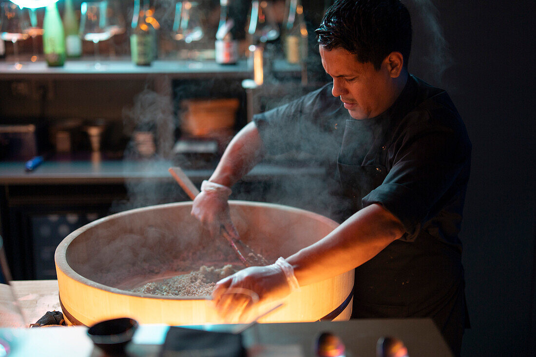 A focused sushi chef prepares fresh rice for sushi dishes in the ambient setting of a sushi restaurant.