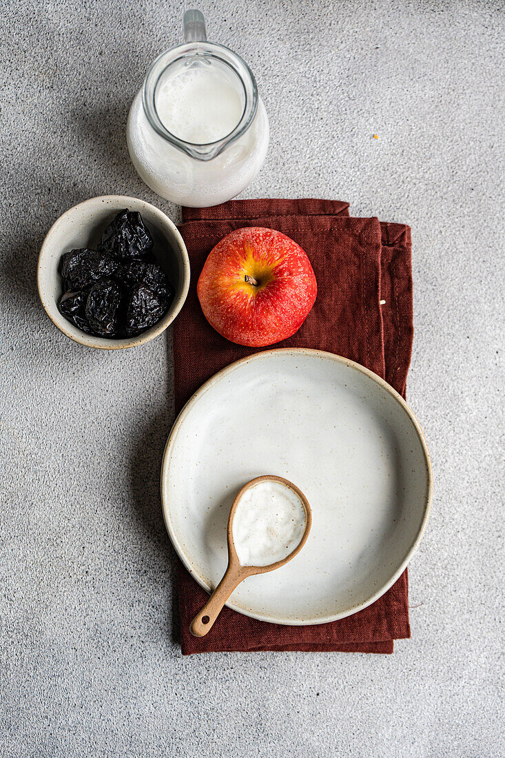 Draufsicht auf eine Schale mit Rosinen auf einem grauen Tisch neben einem leeren weißen Teller und einem Löffel, einem Apfel und einem Milchkännchen