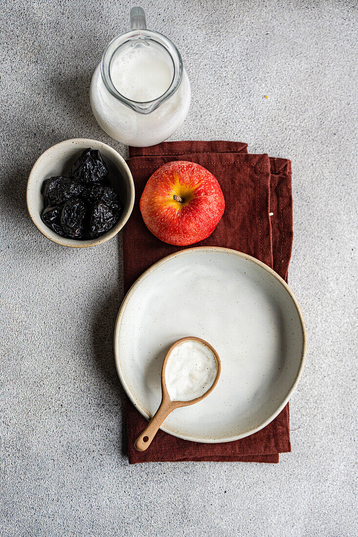 Top view of bowl with raisins placed on gray table near empty white plate and spoon, apple and milk jar