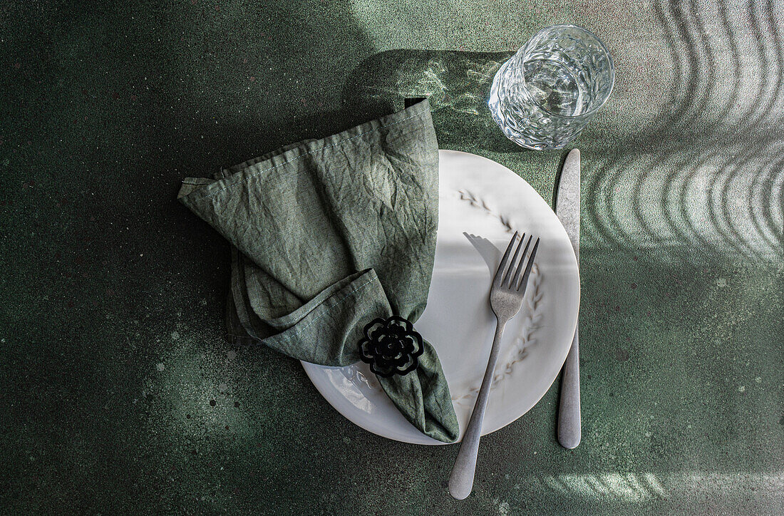 Ein raffiniert gedeckter Tisch mit einem weißen Teller, Besteck, einer Serviette mit Ring und einem Glas, das spielerische Schatten und Reflexionen wirft