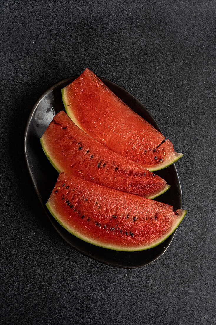 Draufsicht auf Scheiben von Wassermelone, serviert auf schwarzem Teller vor dunkler Oberfläche