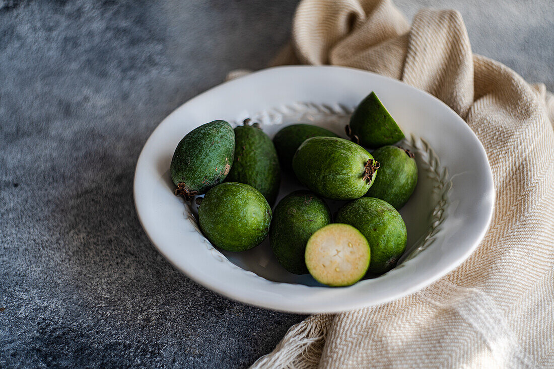 Ripe feijoa fruits arranged neatly in a white ceramic bowl, with a textured cloth background.